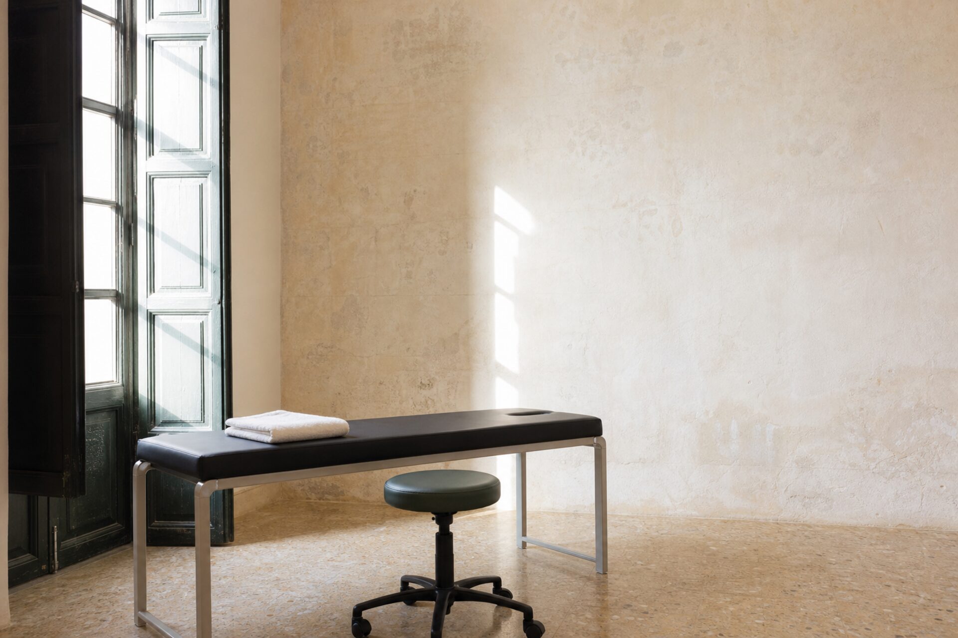 A treatment room at Northgate Physio: tall green louvred shutters open to the street, a black plinth and a stool on a pale terrazzo floor in the morning light.